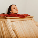 Smiling woman receiving herbal steam therapy in a wooden steam cabinet, head outside with a towel around her neck