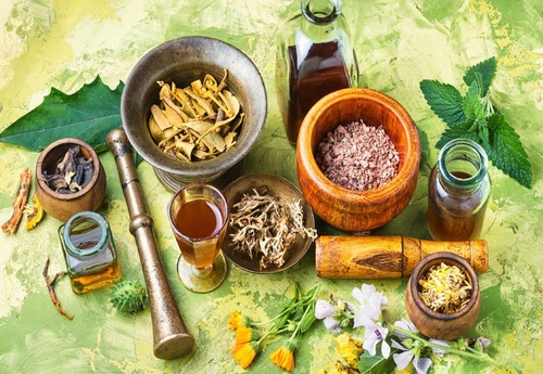 Various herbal medicines, including dried herbs, roots, and oils, displayed in wooden bowls and jars, along with a mortar and pestle on a textured green background.