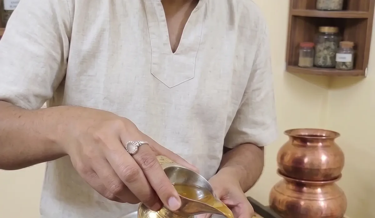 A professional male Ayurvedic therapist in a beige linen kurta performing a Netra Dhara (eye wash) treatment on a male patient lying down. The therapist gently pours a stream of herbal liquid from a traditional brass vessel into the patient's eye