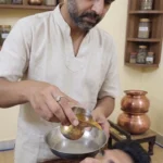 A professional male Ayurvedic therapist in a beige linen kurta performing a Netra Dhara (eye wash) treatment on a male patient lying down. The therapist gently pours a stream of herbal liquid from a traditional brass vessel into the patient's eye