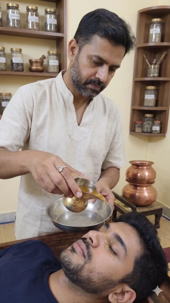 A professional male Ayurvedic therapist in a beige linen kurta performing a Netra Dhara (eye wash) treatment on a male patient lying down. The therapist gently pours a stream of herbal liquid from a traditional brass vessel into the patient's eye